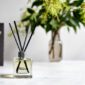 Olieve & olie reed diffuser on a table with vase of flowers behind it. Front view.